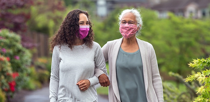 A younger woman walks with an older woman, their arms linked. Both are wearing cloth face masks and smiling.
