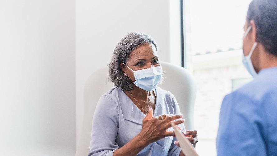 A senior female patient gestures while describing her symptoms to a female healthcare provider. The patient and doctor are wearing protective face coverings. Una paciente mayor hace un gesto mientras describe sus síntomas a una profesional de la salud. El paciente y el médico llevan protectores faciales.