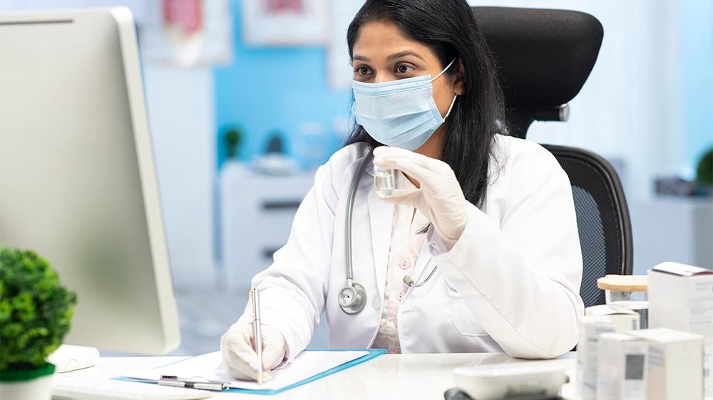 A young doctor sits at her computer in a clinic. She is wearing a face mask and holding a vial. Una joven doctora sentada frente a su computadora en una clínica. Lleva puesta una mascarilla y sostiene un vial.