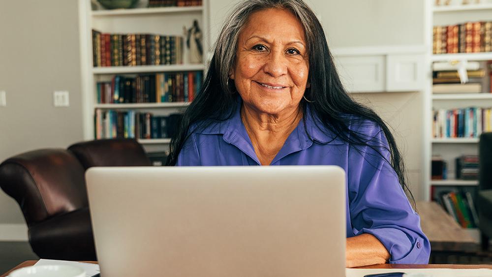 A smiling woman sits at her kitchen table with her laptop. Una mujer sonriente está sentada en la mesa de la cocina con su computadora portátil.