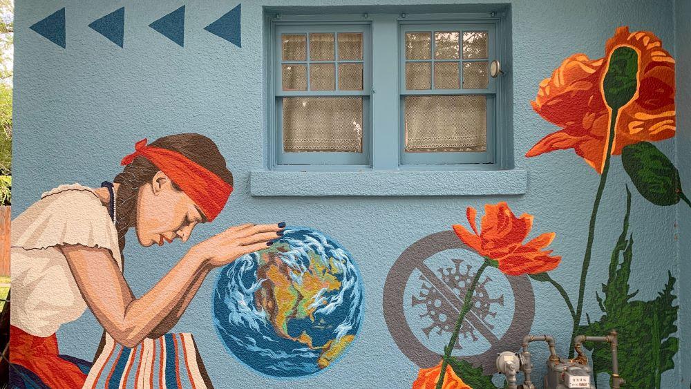 A colorful mural on the side of a blue building. A woman appears to be kneeling in prayer over the earth. There are orange flowers painted around her.