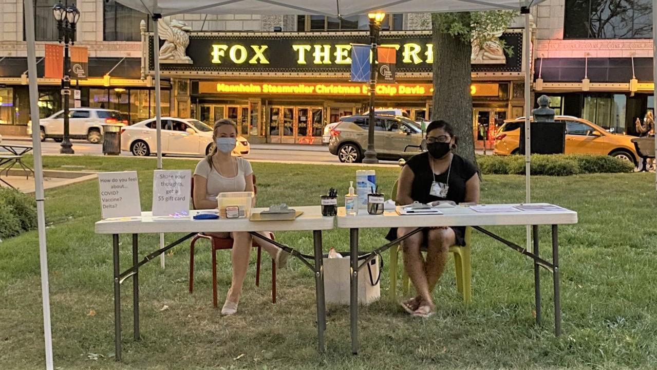 Two women wearing masks sit at a table offering information about COVID-19 and vaccinations