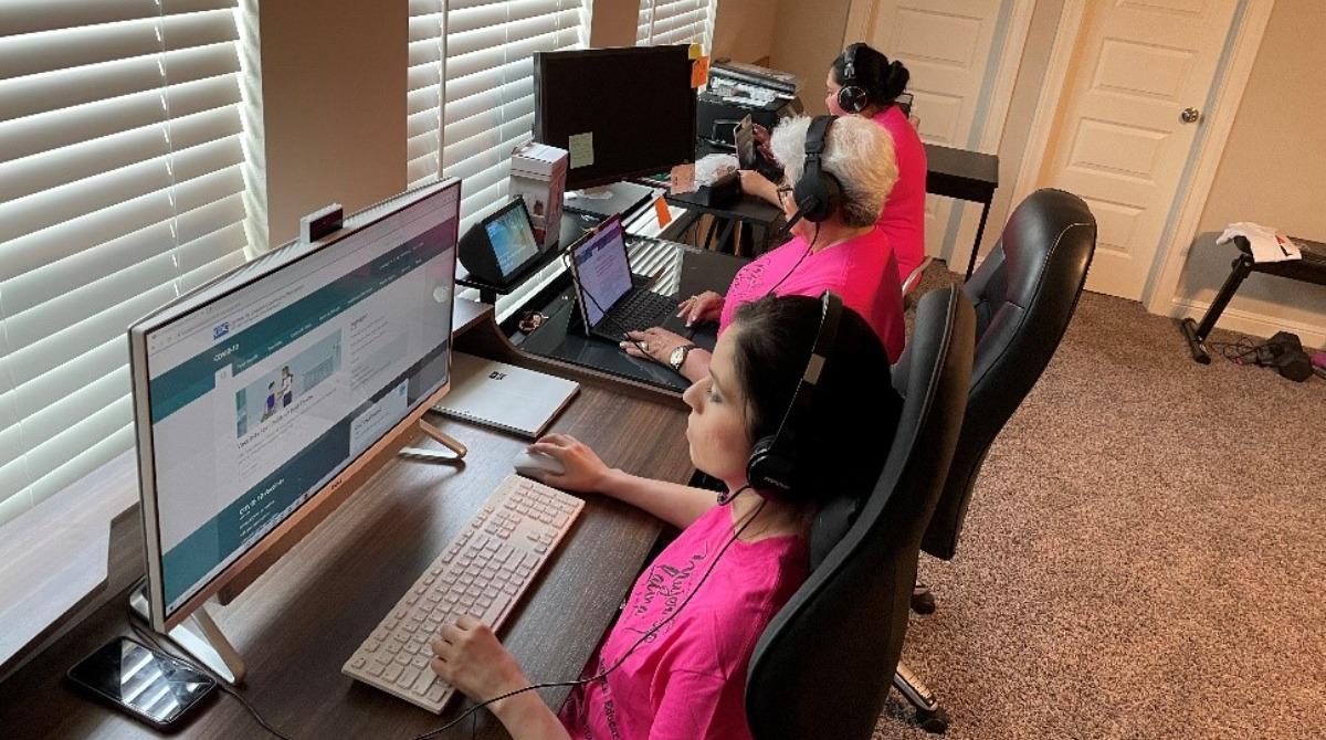 Three women in matching shirts sit at desktop computers. They are part of the Telehealth Community Navigation Center. Tres mujeres con camisas a juego sentadas frente a computadoras de escritorio. Son parte del Centro de Navegación Comunitaria de Telesalud.