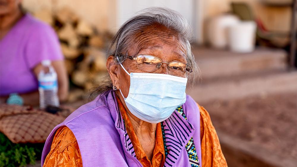 An American Indian woman wearing a mask looks into the distance. Una mujer indígena norteamericana con una mascarilla mira a lo lejos.