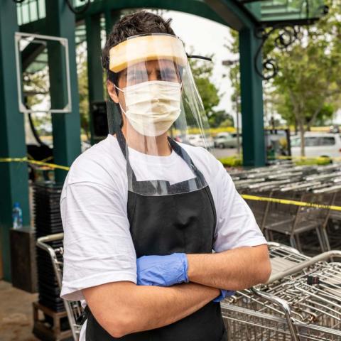 Un hombre con un tapaboca y un protector facial está afuera frente a una tienda de comestibles